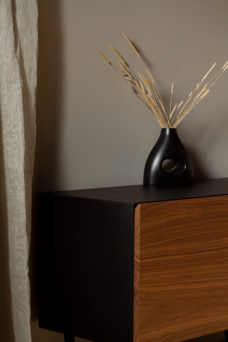 May include: A black wooden cabinet with a single drawer. The drawer is made of wood with a natural grain. The cabinet is sitting on a black surface. A black vase with a cutout design holds dried grasses.