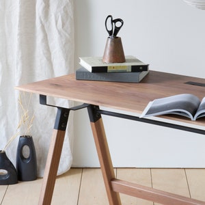 May include: A wooden desk with a light brown tabletop and angled wooden legs. Black metal supports connect the legs to the tabletop. A copper-colored pencil holder with black scissors sits on top of two books. A vase and a decorative vase are on the floor.