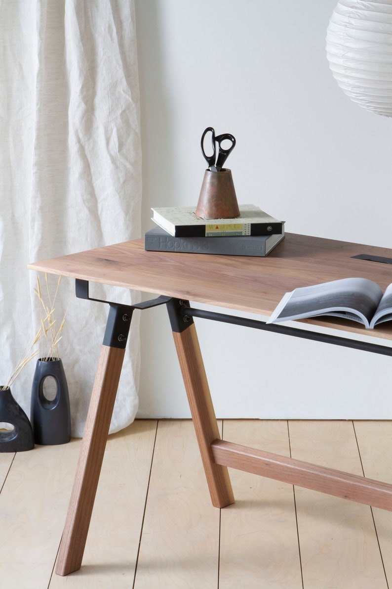 May include: A wooden desk with a light brown tabletop and angled wooden legs. Black metal supports connect the legs to the tabletop. A copper-colored pencil holder with black scissors sits on top of two books. A vase and a decorative vase are on the floor.