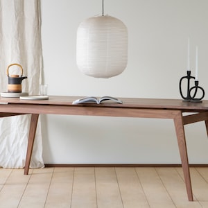 May include: A wooden dining table with a light brown finish and a white paper lantern hanging above it. The table has a rectangular top and four legs with a unique angled design. There are two black candle holders on the table, and a book is lying open on the table.