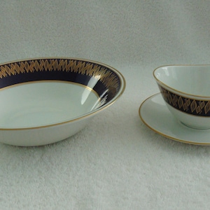 以下が含まれることがあります： 白い磁器のサービングボウルとソーサー付きのティーカップ。どちらの作品にも、縁に金色の葉のデザインが施された濃い青色のバンドがあります。ボウル、ティーカップ、ソーサーの縁は金色のトリムです。