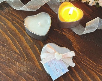 May include: Two heart-shaped candle holders with a white candle in one and a lit candle in the other. A third heart-shaped holder is decorated with a pink ribbon and a tag that says "Benvenuta Camilla 20.12.2023".