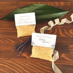 May include: Two small clear bags filled with grains, each with a white label featuring text and dates. A large green leaf and a ribbon are on a dark wooden surface. Dried lavender and a ribbon are also present.