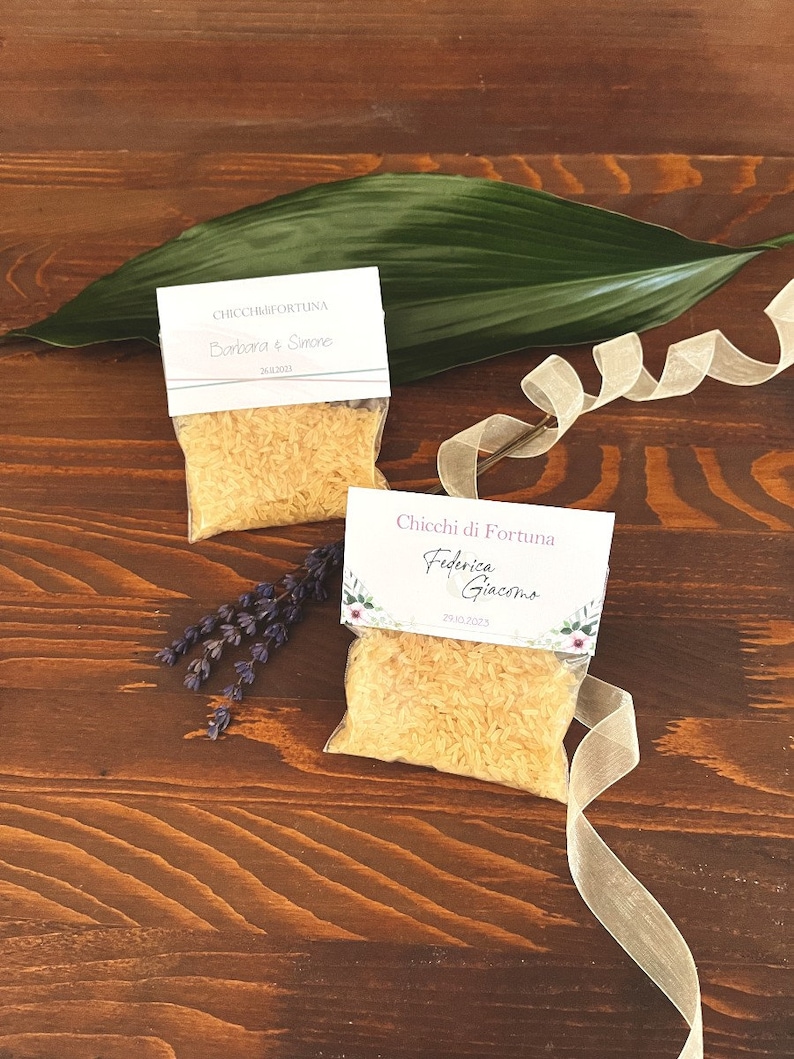 May include: Two small clear bags filled with grains, each with a white label featuring text and dates. A large green leaf and a ribbon are on a dark wooden surface. Dried lavender and a ribbon are also present.
