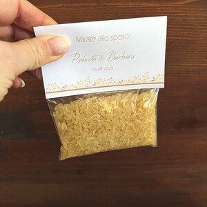 May include: A small clear plastic bag filled with uncooked rice, held by a hand. A white card is attached to the bag, with the text "Mirate allo sposo!" and the names "Roberto & Barbara" along with the date "15.09.2023" in gold lettering.