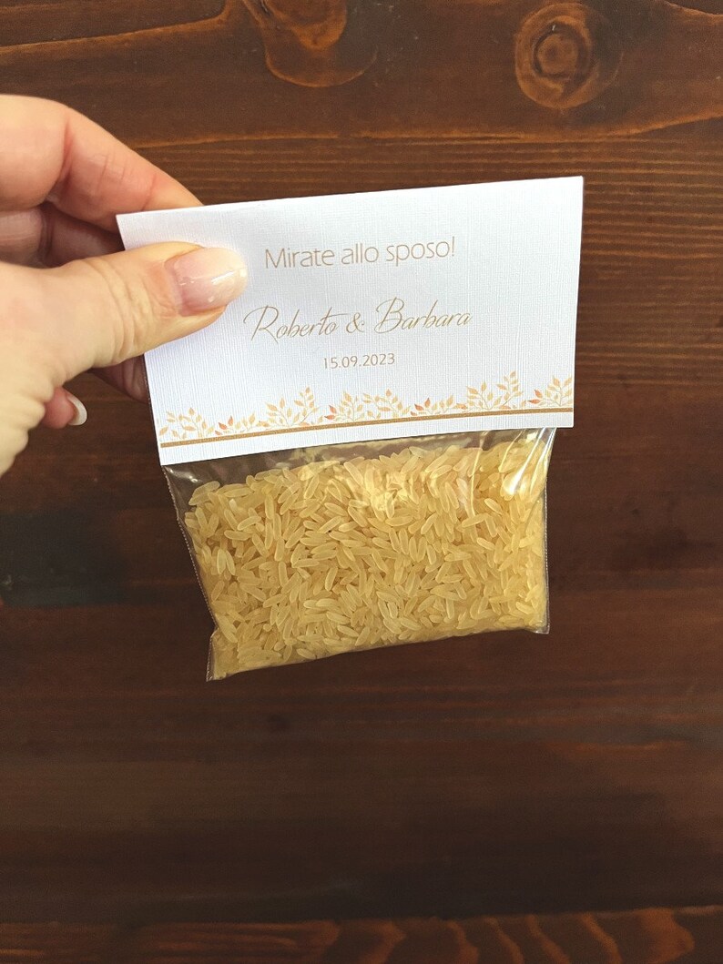 May include: A small clear plastic bag filled with uncooked rice, held by a hand. A white card is attached to the bag, with the text "Mirate allo sposo!" and the names "Roberto & Barbara" along with the date "15.09.2023" in gold lettering.