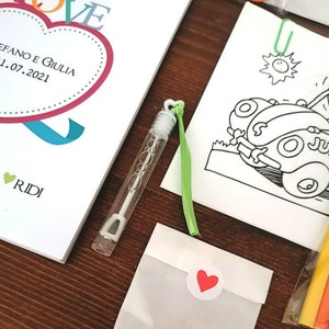 May include: A wedding invitation with a heart design and the text "STEFANO E GIULIA 11.07.2021" in red and black. The invitation is accompanied by a small bottle of bubbles, a green ribbon, a white envelope with a red heart sticker, and a coloring page featuring a cartoon car.