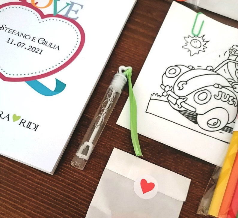 May include: A wedding invitation with a heart design and the text "STEFANO E GIULIA 11.07.2021" in red and black. The invitation is accompanied by a small bottle of bubbles, a green ribbon, a white envelope with a red heart sticker, and a coloring page featuring a cartoon car.
