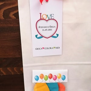 May include: A white paper gift bag with a red ribbon and a tag that says "LOVE" with a heart that says "Stefano e Giulia 11.07.2021" and "GIOCA COLORA RIDI". The bag contains a small plastic bag with three colorful balloons.