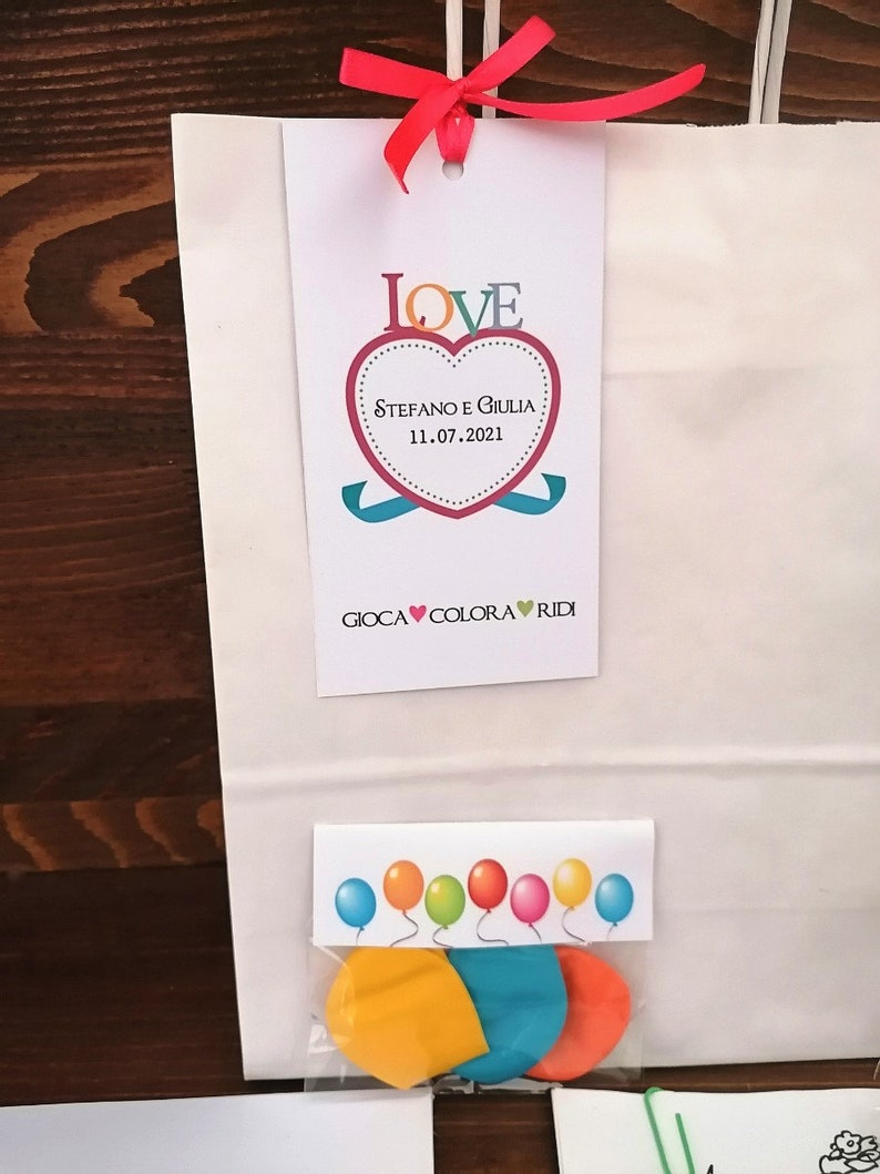 May include: A white paper gift bag with a red ribbon and a tag that says "LOVE" with a heart that says "Stefano e Giulia 11.07.2021" and "GIOCA COLORA RIDI". The bag contains a small plastic bag with three colorful balloons.