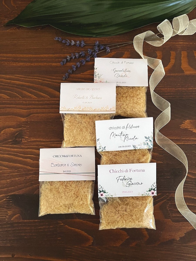 May include: Small clear plastic bags filled with rice, each topped with a decorative card. The cards feature names and dates, with floral and gold accents. The bags are arranged on a dark wooden surface.