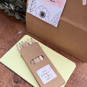 May include: A gift box with a tag that reads "Clarissa - 26.09.2026 - Giacomo". A small box of colored pencils sits on a yellow notepad. The pencils have different colored tips.