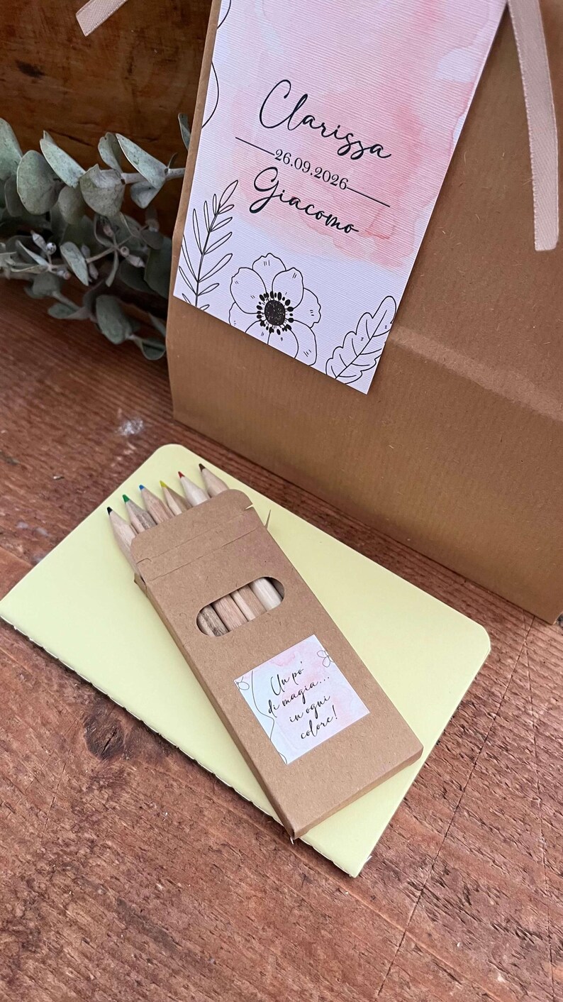 May include: A gift box with a tag that reads "Clarissa - 26.09.2026 - Giacomo". A small box of colored pencils sits on a yellow notepad. The pencils have different colored tips.