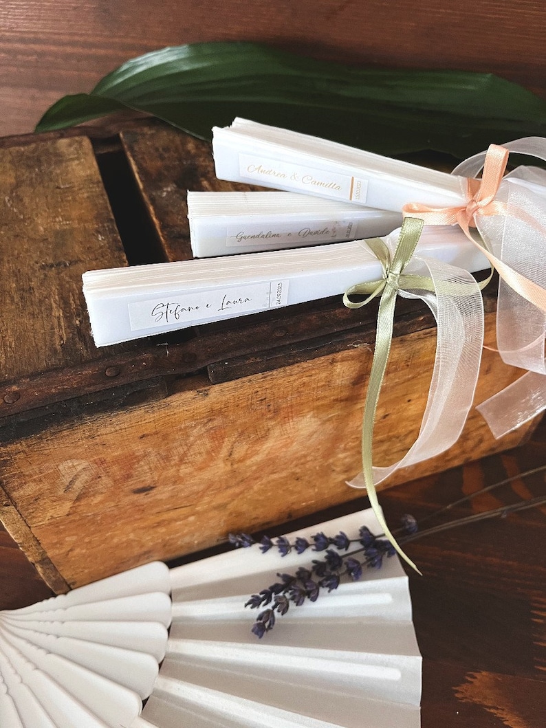 Puede incluir: Tres abanicos de papel blanco con letras doradas atados con una cinta blanca y cintas de color verde y melocot&oacute;n. Los abanicos est&aacute;n etiquetados con "Stefano e Laura", "Giuseppina e Davide" y "Andrea e Camilla".