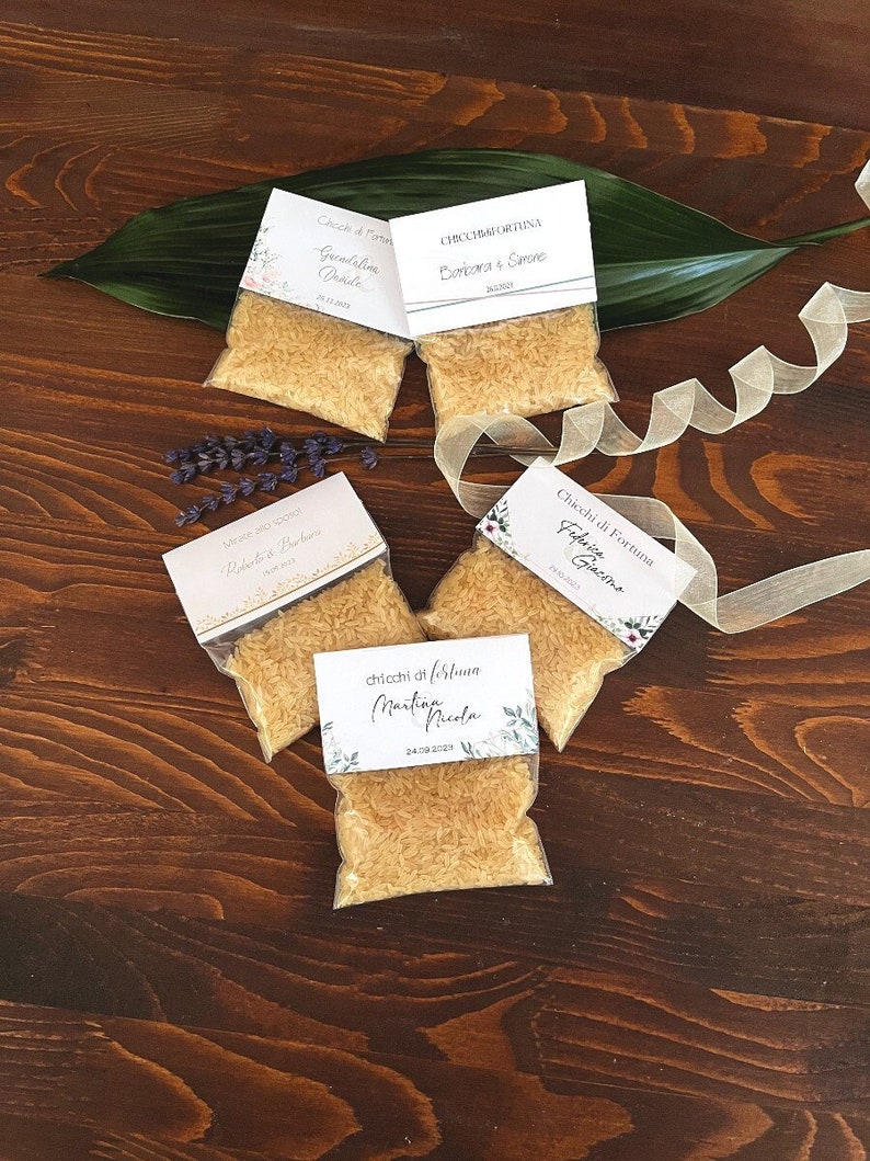 May include: Small clear bags filled with rice, each with a decorative card attached. The cards have floral designs and handwritten text. The bags are arranged on a dark wooden surface with a green leaf and ribbon.