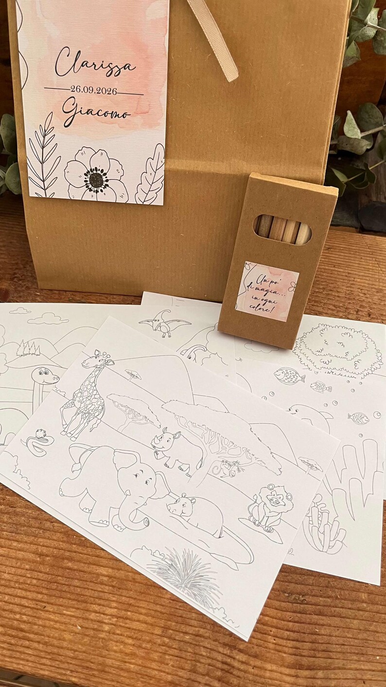 May include: A brown paper gift bag with a floral tag that reads "Clarissa - 26.09.2026 - Giacomo." Several black and white coloring pages with animal illustrations are in front of the bag, along with a small box of colored pencils.