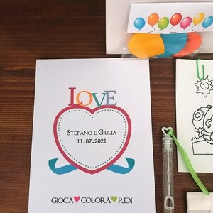 May include: A white card with a red heart that says "LOVE" in colorful letters. The heart has the text "Stefano e Giulia 11.07.2021" inside. Below the heart is the text "GIOCA &hearts; COLORA &hearts; RIDI".
