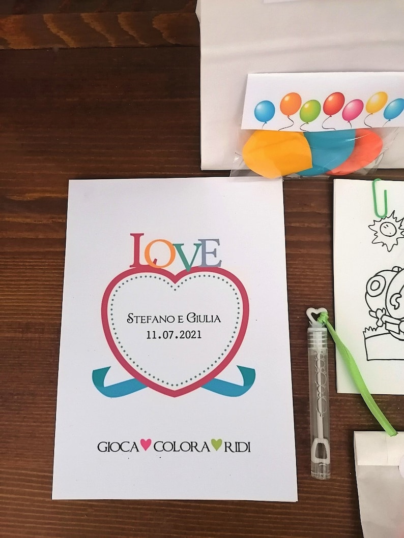 May include: A white card with a red heart that says "LOVE" in colorful letters. The heart has the text "Stefano e Giulia 11.07.2021" inside. Below the heart is the text "GIOCA &hearts; COLORA &hearts; RIDI".