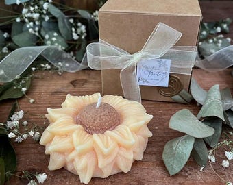 May include: A sunflower-shaped candle with a brown center and yellow petals sits on a wooden surface. A brown box tied with a sheer ribbon and a sprig of greenery are in the background. The candle is a decorative item.