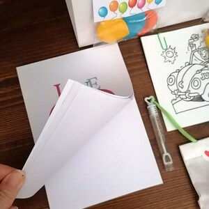 May include: A white card with the word "Love" written in pink, blue, and green letters. The card is folded in half and is sitting on a wooden surface. There are other items in the image, including a bag, a coloring page, and a bottle of bubbles.