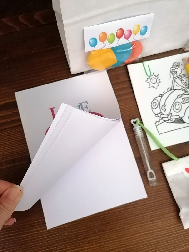 May include: A white card with the word "Love" written in pink, blue, and green letters. The card is folded in half and is sitting on a wooden surface. There are other items in the image, including a bag, a coloring page, and a bottle of bubbles.