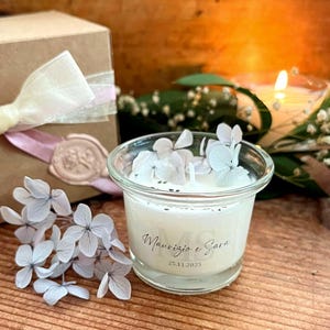 Può includere: Un piccolo portacandele in vetro trasparente con una candela bianca, decorata con piccoli fiori bianchi. La candela riporta i nomi "Maurizio e Sara" e la data "25.11.2025". Sullo sfondo, una scatola regalo con un nastro e un fiore.