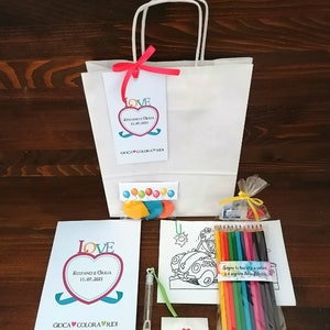May include: A white gift bag with a red ribbon and a tag that says "LOVE STEFANO E GIULIA 11.07.2021 GIOCA COLORA RIDI". The bag contains a coloring book, crayons, a bubble wand, and a small envelope with a heart.
