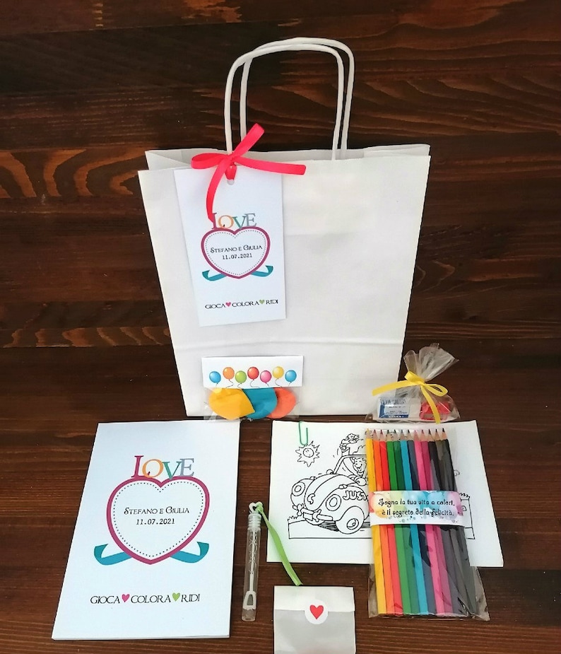 May include: A white gift bag with a red ribbon and a tag that says "LOVE STEFANO E GIULIA 11.07.2021 GIOCA COLORA RIDI". The bag contains a coloring book, crayons, a bubble wand, and a small envelope with a heart.