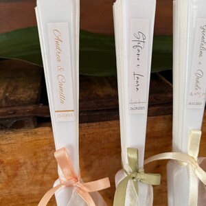 Puede incluir: Tres abanicos plegables blancos con etiquetas personalizadas y cintas. Las etiquetas dicen "Andrea & Camilla 15.10.2023", "Stefano e Lavinia 24.09.2023" y "Giuseppina e Davide 28.11.2023".