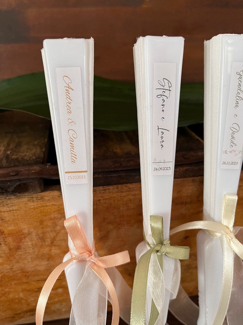 Puede incluir: Tres abanicos plegables blancos con etiquetas personalizadas y cintas. Las etiquetas dicen "Andrea & Camilla 15.10.2023", "Stefano e Lavinia 24.09.2023" y "Giuseppina e Davide 28.11.2023".