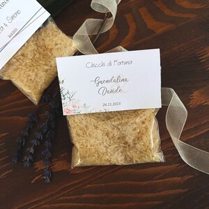 May include: Two small clear plastic bags filled with rice, each with a decorative card attached. The cards have floral designs and text that reads "Chicchi di Fortuna" and names. A ribbon and sprig of lavender are also present.