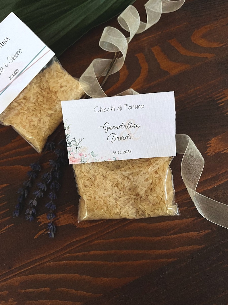 May include: Two small clear plastic bags filled with rice, each with a decorative card attached. The cards have floral designs and text that reads "Chicchi di Fortuna" and names. A ribbon and sprig of lavender are also present.