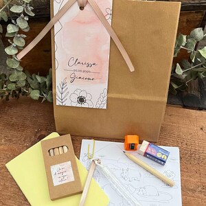 May include: A brown paper gift bag with a pink ribbon and tag, containing art supplies. The tag reads "Clarissa 26.09.2026 Giacomo" with floral illustrations. Includes crayons, a coloring page, and a pencil.
