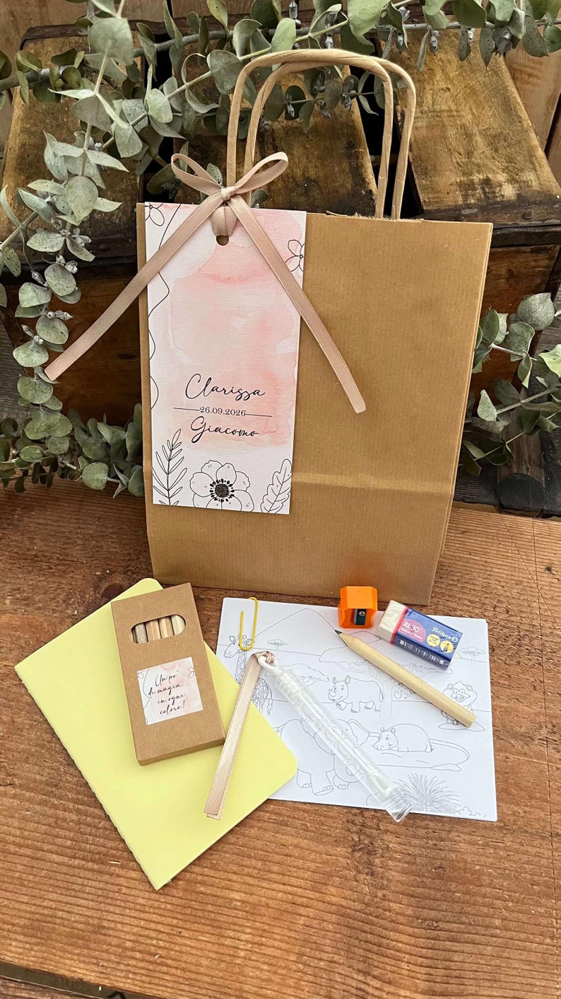 May include: A brown paper gift bag with a pink ribbon and tag, containing art supplies. The tag reads "Clarissa 26.09.2026 Giacomo" with floral illustrations. Includes crayons, a coloring page, and a pencil.