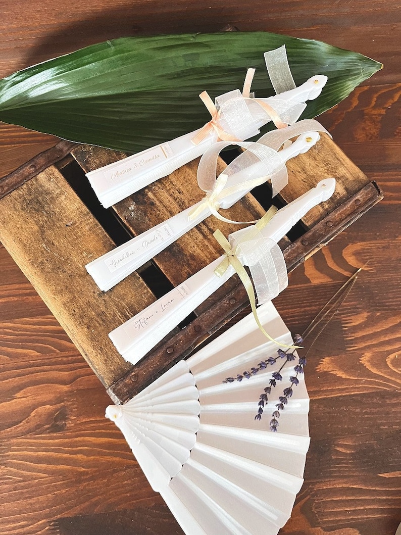Puede incluir: Tres abanicos de papel blanco con cintas decorativas y los nombres de la pareja de bodas impresos en ellos. Los abanicos est&aacute;n dispuestos en una caja de madera con una hoja verde en la parte superior. Un abanico plegable blanco con flores de lavanda est&aacute; en primer plano.