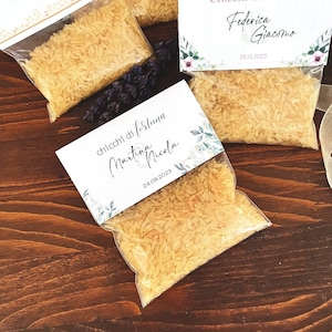 May include: Several small clear plastic bags filled with rice, each topped with a decorative card. The cards feature names and dates, suggesting they are wedding favors. The bags are arranged on a dark wooden surface.