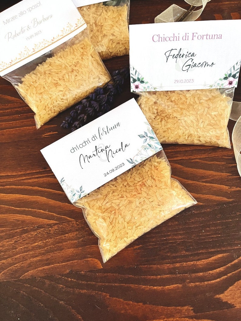 May include: Several small clear plastic bags filled with rice, each topped with a decorative card. The cards feature names and dates, suggesting they are wedding favors. The bags are arranged on a dark wooden surface.