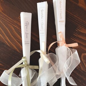 Puede incluir: Tres abanicos de papel blanco con cintas decorativas. Los abanicos tienen etiquetas con los nombres de las parejas y sus fechas de boda: "Stefanie & Laura 24.09.2023", "Gwendaline & David 26.11.2023" y "Andrea & Camilla 13.10.2023".