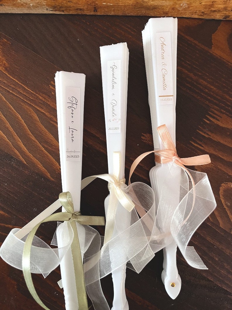 Puede incluir: Tres abanicos de papel blanco con cintas decorativas. Los abanicos tienen etiquetas con los nombres de las parejas y sus fechas de boda: "Stefanie & Laura 24.09.2023", "Gwendaline & David 26.11.2023" y "Andrea & Camilla 13.10.2023".