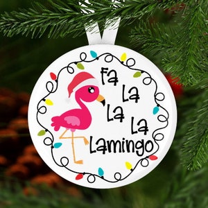 Może przedstawiać: Biała ozdoba świąteczna z r&oacute;żowym flamingiem w czapce Świętego Mikołaja. Ozdoba zawiera napis "Fa La La La Lamingo" i jest ozdobiona łańcuchem kolorowych światełek. Ozdoba wisi na białej wstążce.