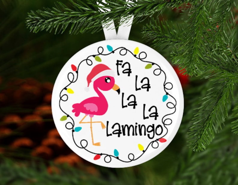 Może przedstawiać: Biała ozdoba świąteczna z r&oacute;żowym flamingiem w czapce Świętego Mikołaja. Ozdoba zawiera napis "Fa La La La Lamingo" i jest ozdobiona łańcuchem kolorowych światełek. Ozdoba wisi na białej wstążce.