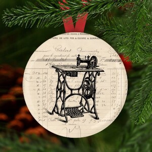 May include: A vintage-style Christmas ornament featuring a black and white illustration of a sewing machine on a white background with black text. The ornament is round and has a red ribbon attached.