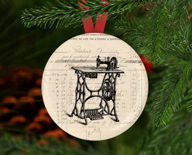 May include: A vintage-style Christmas ornament featuring a black and white illustration of a sewing machine on a white background with black text. The ornament is round and has a red ribbon attached.