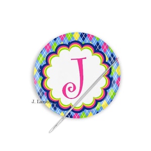 May include: A round, decorative plate with a white center featuring a pink letter "J". The plate has a scalloped edge and is surrounded by a colorful argyle pattern in blue, green, yellow, and pink. A silver needle is placed diagonally across the plate.