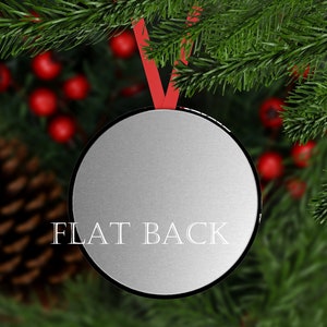 May include: A silver round ornament with a flat back. The ornament is hanging from a red ribbon and is surrounded by a blurry background of green pine branches and red berries.