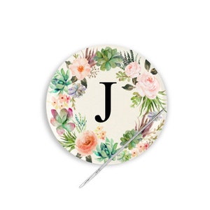 May include: A round floral wreath with a watercolor design featuring pink, green, and white flowers and greenery. The letter "J" is in the center of the wreath.