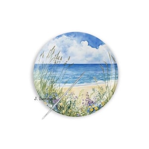 May include: A round decorative plate featuring a watercolor painting of a coastal scene with a sandy beach, blue ocean, and white clouds. Tall grasses and small purple and yellow wildflowers grow in the foreground. The text J. Larson's is visible.