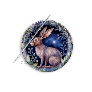 May include: A round, watercolor illustration of a hare inside a floral wreath, with a needle piercing through the center. The hare is brown and tan, with long ears and a watchful gaze. The wreath features blue and green foliage against a dark blue background. Text reads "J. Larson".