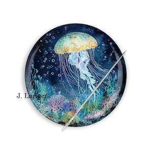 May include: A round needle minder with a watercolor illustration of a jellyfish in a dark blue ocean with coral and bubbles. The needle minder has a silver needle attached to it.