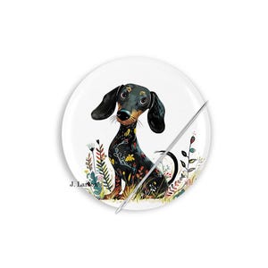 May include: A white round button with a colorful illustration of a dachshund dog. The dog is black with floral patterns and is sitting in a field of flowers. A silver pin is attached to the button.